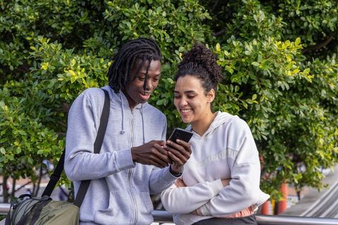 African american young couple sharing smartphone laughing outdoors urban greenery casual