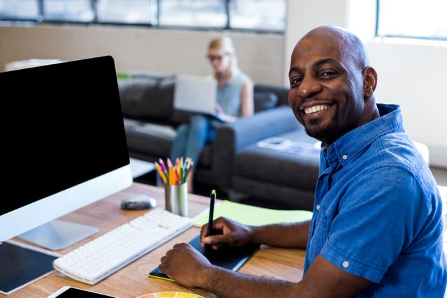Man sitting at his desk in a modern office, smiling at the camera. He is using a computer and has various office supplies around him. In the background, a woman is sitting on a couch working on a laptop. This image can be used for business, technology, and workplace-related content, showcasing a positive and productive work environment.