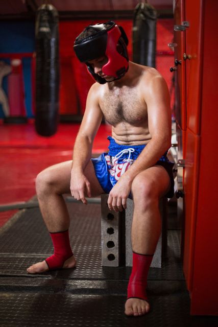 Boxer sitting in locker room after intense training session, wearing boxing gear and helmet. Ideal for use in fitness, sports, and health-related content, emphasizing themes of hard work, dedication, and recovery.