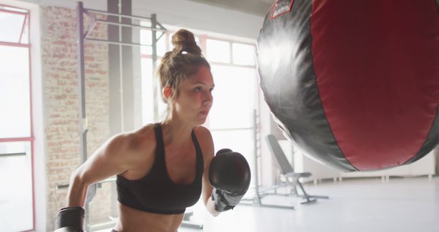 Young woman in fitness attire engaging in boxing workout with punching bag indoors. Ideal for advertising gym memberships, sport equipment, active lifestyle promotions, fitness magazines, and motivation blogs.