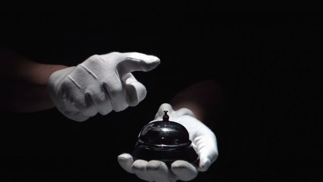 The video depicts gloved hands ringing a hotel bell against a dark background, exuding luxury and elegance. This visual can be used in marketing materials for hotels, luxury service promotions, hospitality industry advertisements, or website banners.