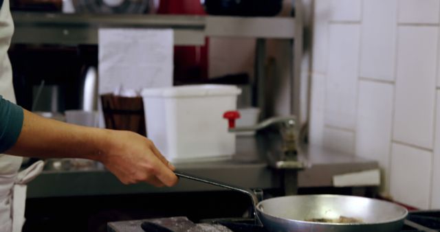 Chef's hand stirring food in a frying pan on a stove in a commercial kitchen. Ideal for use in articles and blogs about culinary arts, restaurant industry insights, cooking techniques, kitchen management, chef career tips, and professional cooking recipes.