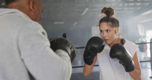 Female boxer sparring with her coach in a gym, preparing for a match. Her focused expression and stance suggest determination and hard work, making this suitable for content related to fitness, combat sports, women's empowerment, and training programs.
