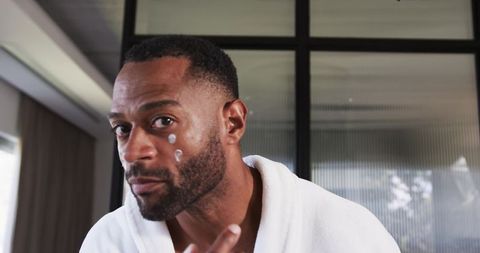 Mature man applying facial cream in white robe near mirror in minimalist bathroom