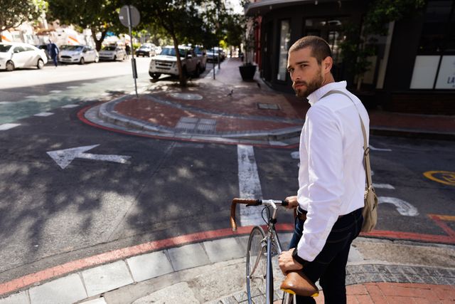 Side view of a handsome elegant Caucasian businessman wearing white shirt holding his bike and looking at camera on a urban street. Digital Nomad on the go.