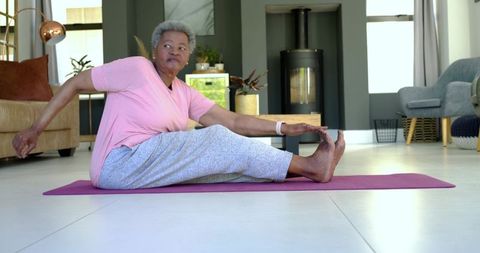 Senior african american woman stretching on purple yoga mat in bright living room