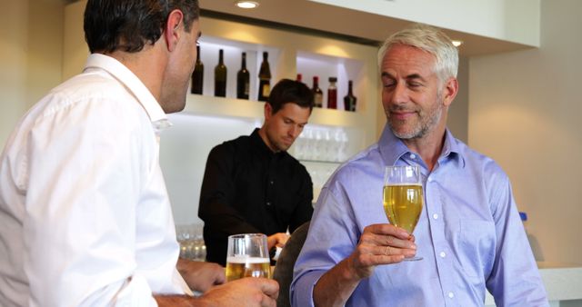 Two men are drinking beer and conversing at bar counter while bartender prepares drinks in background. This could be used for advertisements related to bars, pub culture, social gatherings, or promoting alcoholic beverages and casual leisure activities.