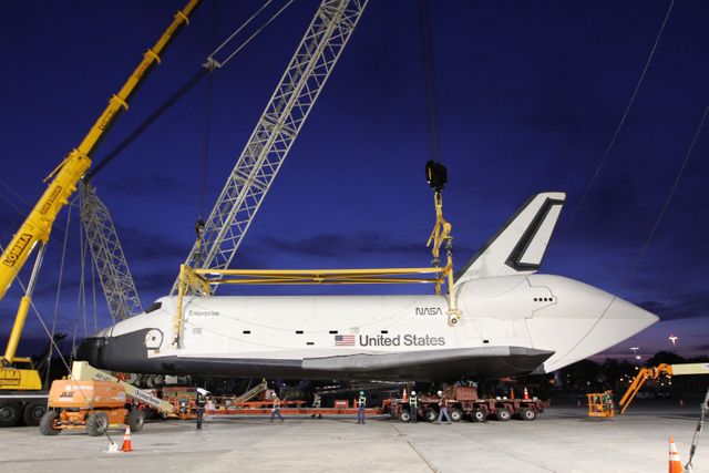 This photo shows the NASA Space Shuttle Enterprise being carefully lowered onto a specialized truck bed using cranes at John F. Kennedy International Airport in New York City. Enterprise is a prototype built to test various aspects of the space shuttle design and is being prepared for transportation to the Intrepid Sea, Air, and Space Museum. The process is taking place during the evening, with cranes, machinery, and crew visible ensuring the safe move of the spacecraft. Ideal for articles or presentations about aerospace history, space exploration, NASA missions, and museum exhibits.