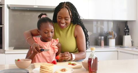 African american mom teaching daughter to slice strawberries for pancakes in modern kitchen