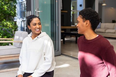Multicultural women chatting on sunlit patio near open sliding door wearing hoodie and knit sweater