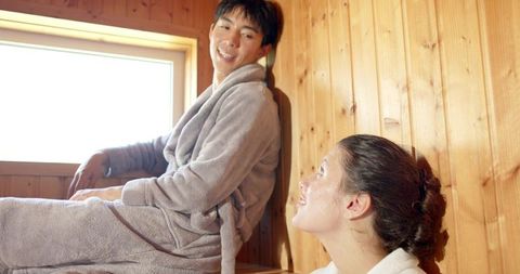 Sunlit spa sauna couple relaxing in wooden sauna wearing robes, asian man caucasian woman