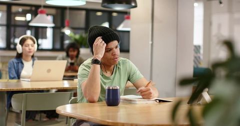 Young man studying in coworking cafe wearing beanie writing in notebook with tumbler and tablet