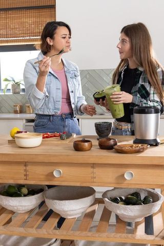 Female friends preparing and tasting green avocado smoothie in bright home kitchen