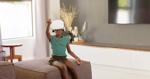 African american teen girl sitting on ottoman using white vr headset in bright living room