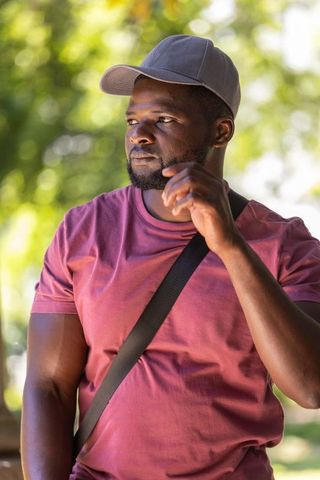 African man adjusting cap in sunlit park wearing maroon tee and crossbody strap