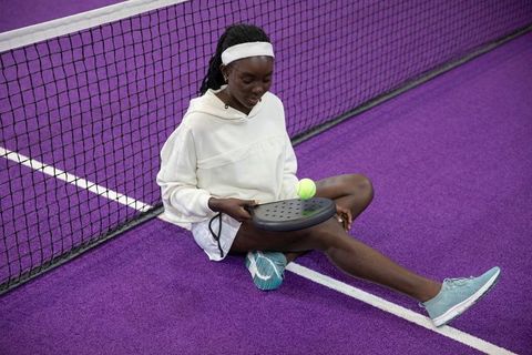 African american woman sitting on purple court holding pickleball paddle and ball near net