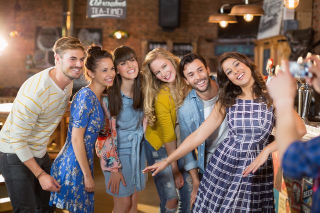 Group of happy friends posing while man photographing through mobile phone in pub