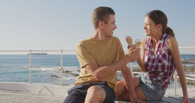 Young couple delighting in cold ice cream near the ocean on a clear summer day. Ideal for promoting summer vacations, romance, seaside activities, and joyful moments.