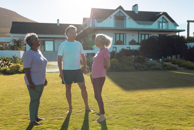 Senior multiracial friends talking while standing on grassy land in front of nursing home in yard. Summer, unaltered, togetherness, support, assisted living and retirement concept.