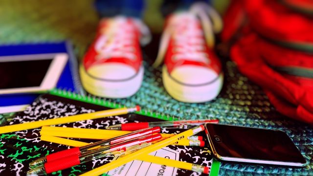 Colorful layout of school supplies including notebooks, pencils, and pens spread out on a blue-green mat. Red sneakers and a red backpack are visible in the background. Suitable for use in educational materials, advertisements for back-to-school sales, or student-oriented content.