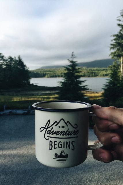 Hand holding adventure-themed mug with the phrase 'The Adventure Begins' in front of scenic mountain and forest landscape. Suitable for use in travel blog articles, motivational content, and promotional material for outdoor activities and adventures.