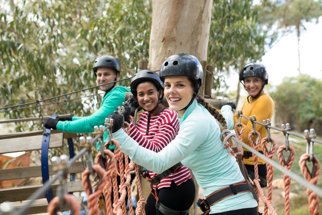 Group of friends standing on a rope bridge in an adventure park, wearing helmets and safety gear. They are smiling and appear to be enjoying the outdoor activity. This image can be used for promoting adventure parks, team-building activities, outdoor fun, and group bonding experiences.
