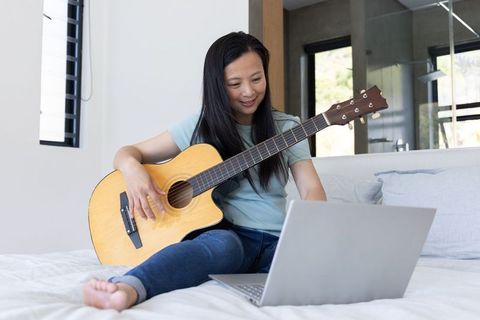 Mature asian woman practicing acoustic guitar on bed while using laptop in modern bedroom