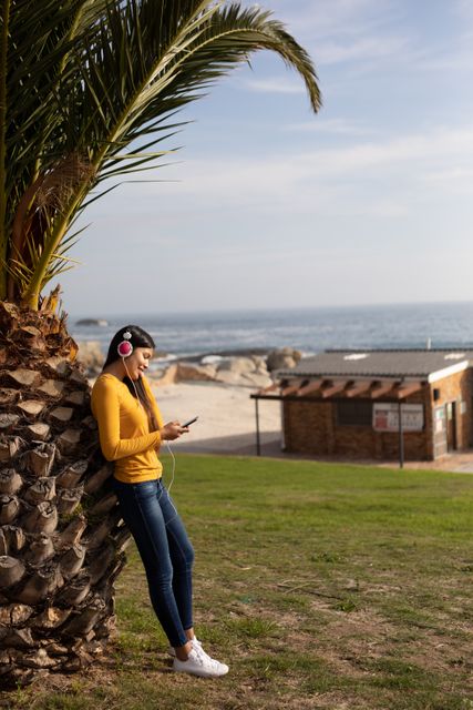 Young biracial woman leaning against a palm tree, smiling while using a smartphone and wearing headphones on a sunny day. Ideal for concepts related to leisure, technology, connectivity, modern lifestyle, and enjoying free time outdoors. Perfect for advertisements, blogs, and articles about digital devices, music, and relaxation in nature.