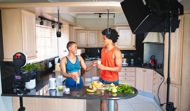 Two friends are in a modern kitchen, discussing and vlogging about healthy food. They are surrounded by fresh fruits and vegetables, and there is a camera setup indicating content creation for social media. This image is perfect for promoting healthy lifestyles, fitness blogs, cooking channels, and social media content related to nutrition and wellness.