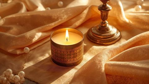 Glowing amber candle in ornate brass tin on peach-gold satin with warm ambient glow