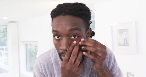 African American man removing contact lens in bathroom mirror during morning grooming routine