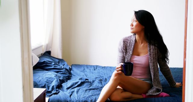 A woman is sitting on her bed with a blue duvet, holding a cup of coffee and looking out of the window. The scene conveys a sense of relaxation and peaceful morning routine. This photo can be used for lifestyle blogs, advertisements for home goods, and articles about self-care or morning routines.