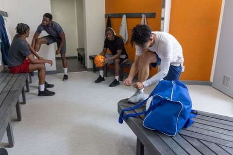 Multicultural basketball teammates preparing in locker room while asian player tying shoe