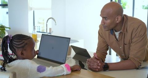 Father guiding daughter using laptop and tablet in bright kitchen during home learning