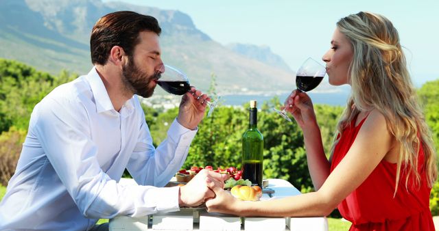 A Caucasian couple enjoys a romantic outdoor meal with a scenic mountain backdrop, with copy space. They are toasting with glasses of red wine, enhancing the intimate atmosphere of their date.
