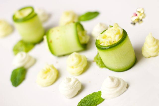 This visually appealing composition shows gourmet zucchini rolls filled with cream cheese, garnished with fresh mint leaves and dabs of cream, plated elegantly on a white surface. Ideal for use in culinary magazines, restaurant menus, food blogs, or promotional materials for gourmet food events.