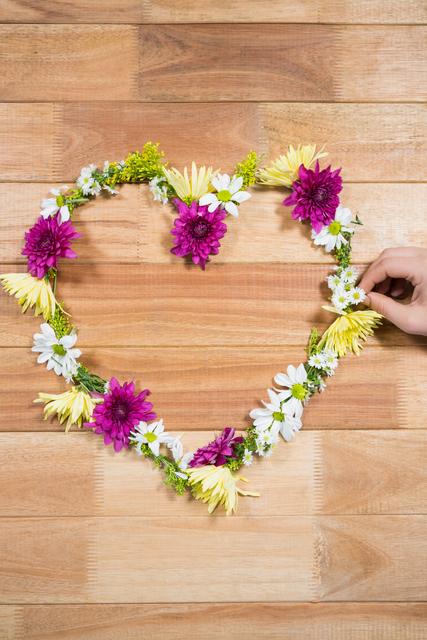 Hand arranging a tropical flower garland in a heart shape on a wooden board. Ideal for use in DIY projects, crafting tutorials, romantic decorations, wedding planning, and nature-inspired designs. Perfect for blogs, social media posts, and websites focused on creativity, love, and handmade crafts.