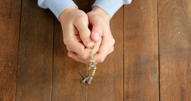 A child's hands are clasped in prayer, holding a rosary with a wooden background, with copy space. It's a moment of spirituality or religious practice, reflecting faith and devotion.