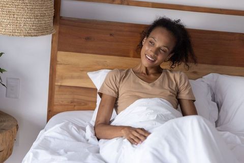 African american woman relaxing in bed embracing white comforter in boho bedroom morning