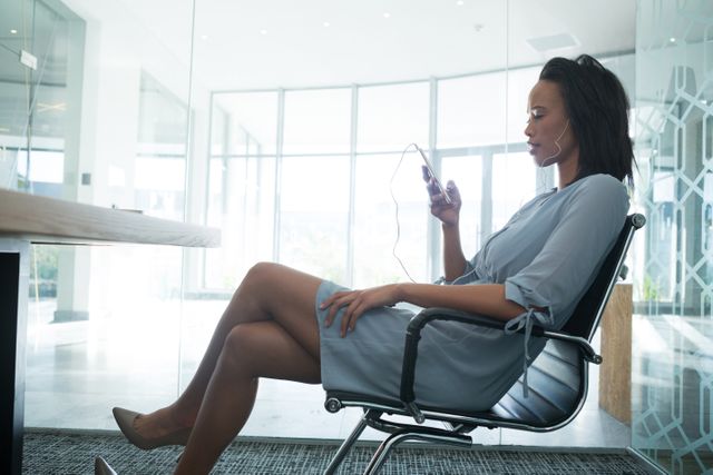 Female executive in business attire sitting in a modern office, listening to music on her mobile phone with headphones. Ideal for use in articles or advertisements related to workplace relaxation, modern office environments, corporate culture, and technology in the workplace.