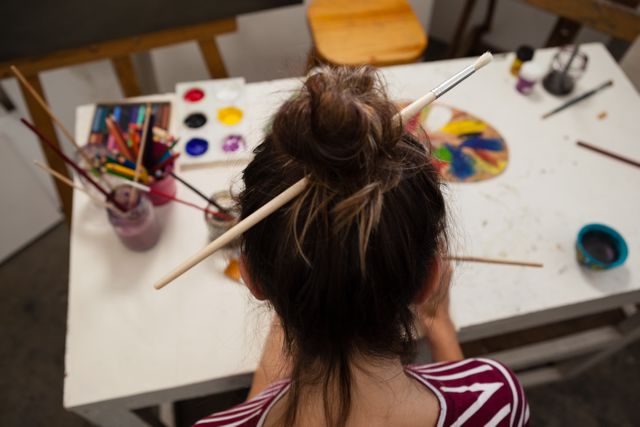 Woman painting in an art class, seen from behind, with paintbrushes in her hair and a palette on the table. Ideal for use in articles about art education, creativity, hobbies, and artistic expression.