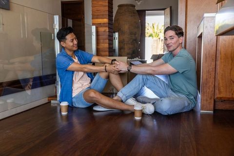 Diverse friends sitting on hardwood floor holding hands sharing coffee in modern foyer