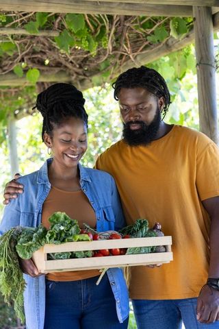 African couple holding crate of fresh garden vegetables beneath wooden pergola, smiling