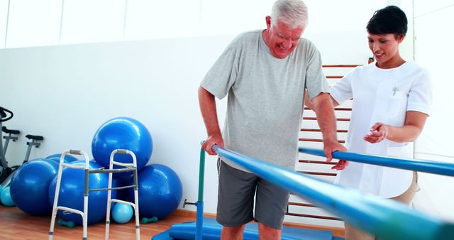 This image shows an elderly man engaging in a physical therapy session with the support of a nurse. The nurse assists the man as he holds onto parallel bars for stability. There are various rehabilitation equipment, including exercise balls and a walker, visible in the background. Ideal for use in articles or educational material highlighting elderly care, rehabilitation, physical therapy sessions, and the importance of healthcare support for senior citizens.