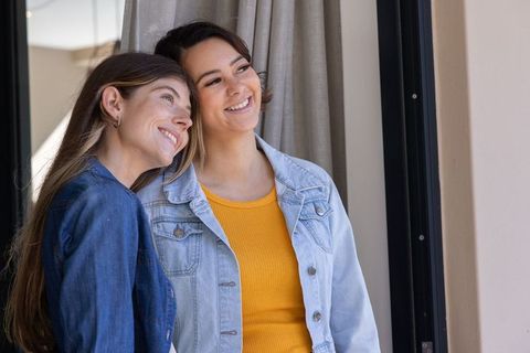 Two women smiling and leaning together in denim jackets with mustard top sharing warm moment at home
