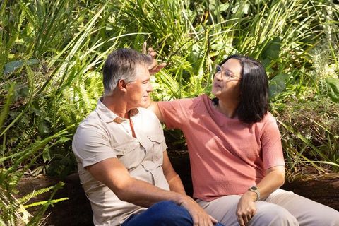 Diverse mature couple relaxing on log in sunlit park surrounded by ferns