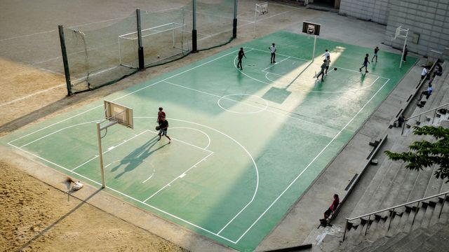 Shot showcasing a group of young players engaged in a basketball game on an outdoor court under daylight. Useful for depicting outdoor sports activities, teamwork, youth fitness, and recreational activities. Ideal for advertisements related to sports, fitness clubs, and youth programs.
