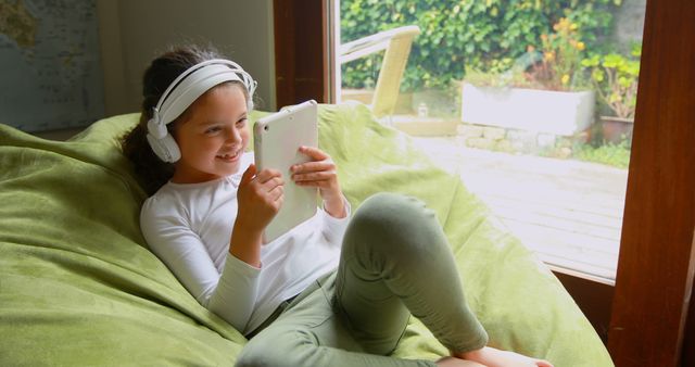A young girl is smiling and wearing headphones while using a tablet. She is reclining on a green bean bag in a cozy home environment. The bright light from the windows in the background contrasts with the indoor scene, where the child appears to be enjoying digital music or a video. This image is ideal for use in advertising children’s products, family lifestyle blogs, technology-related content, or educational materials focused on digital learning and entertainment.