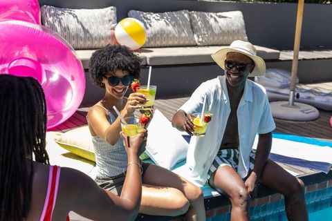 Diverse friends clinking cocktails poolside, dipping feet, smiling in swimwear, sunglasses