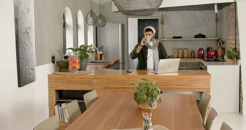 Indian man working remotely in contemporary kitchen, sipping coffee while using laptop